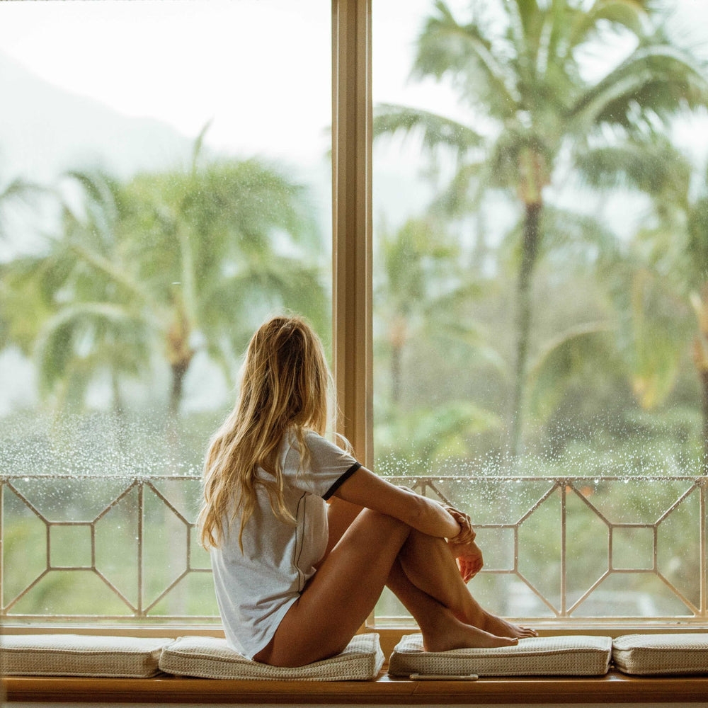 Frau in weißen Tshirt schaut aus Fensterfront in Natur mit Palmen
