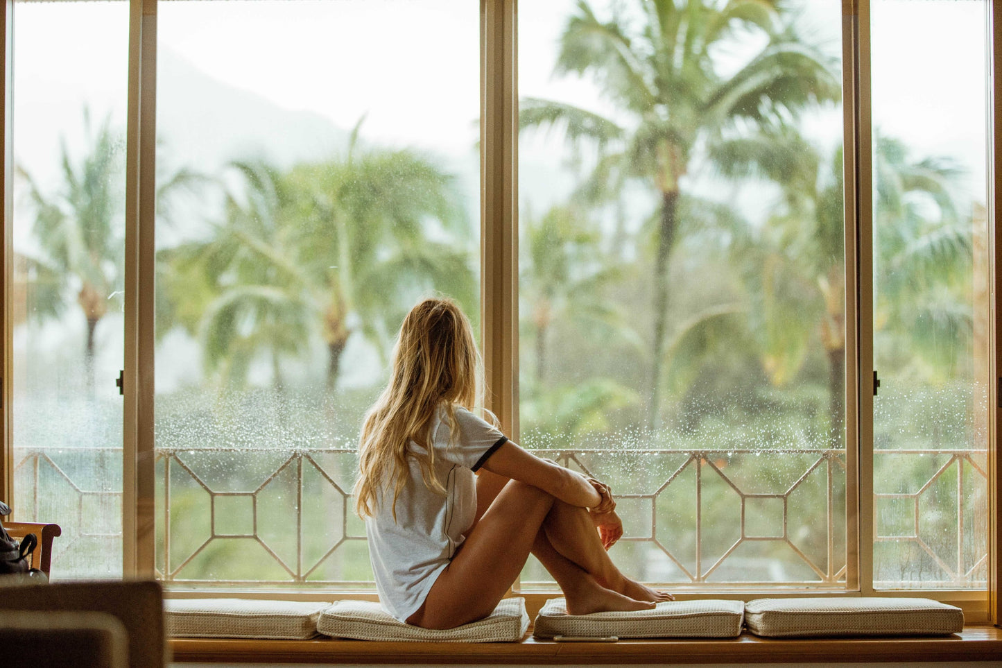 Frau in weißen Tshirt schaut aus Fensterfront in Natur mit Palmen