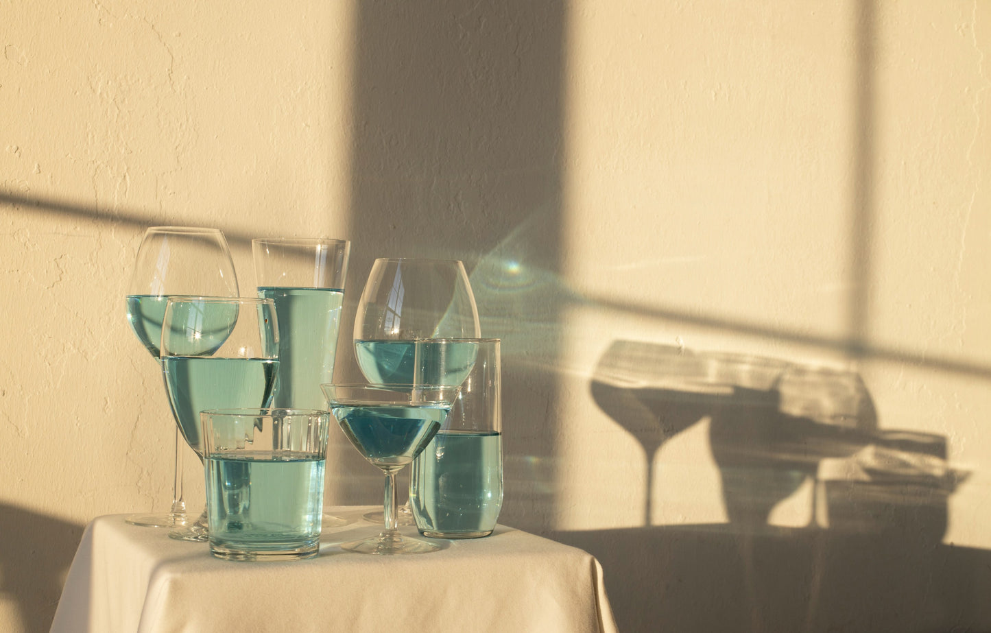 Gläser mit hellblauer durchsichtiger Flüssigkeit auf Tisch in Sonne mit Schatten auf gelblicher Wand 
