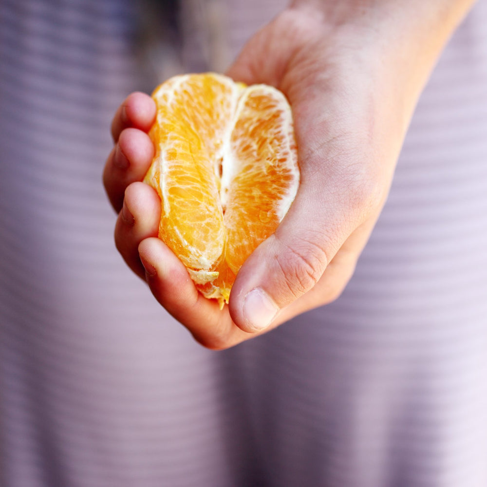 Frauenhand mit Orange in Form wie Vulva in Hand vor lila Hintergrund